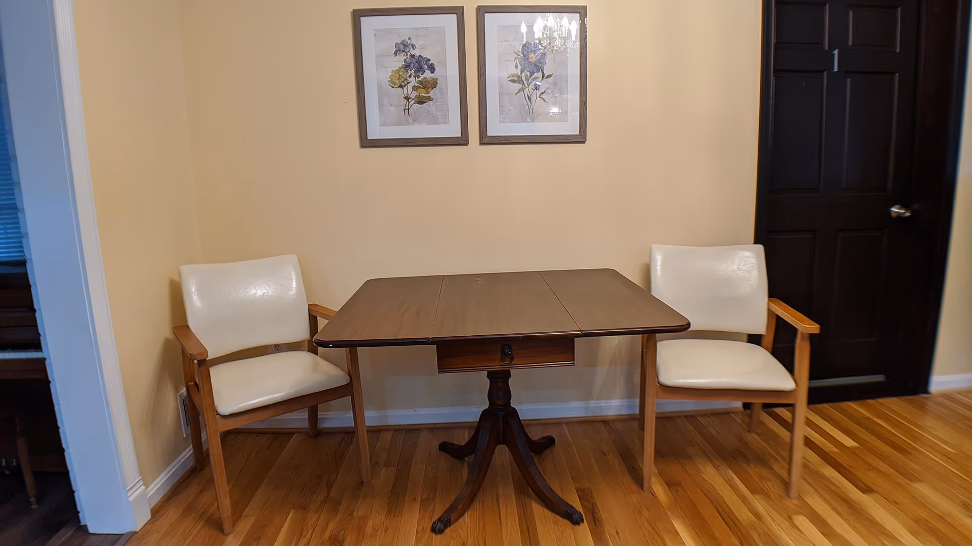 A small wooden table with a central pedestal base and two white cushioned wooden chairs on either side, placed against a beige wall with two framed floral pictures hanging above. The floor is hardwood, and there is a dark brown door with the number 1 on it to the right.