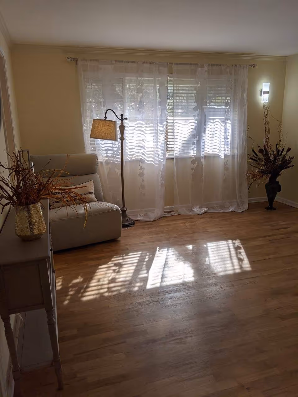 Sunlit living room with sheer curtains over a large window, a floor lamp next to an armchair, a small side table with a vase, and hardwood flooring.