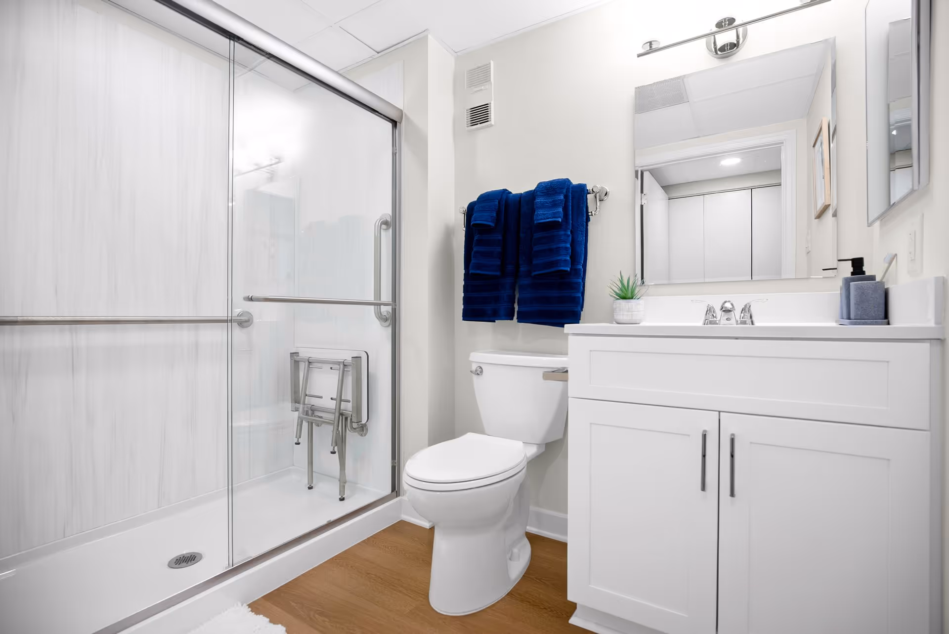 Bright modern bathroom with a glass-enclosed shower, toilet, white vanity and mirror, and blue towels hanging on the wall.