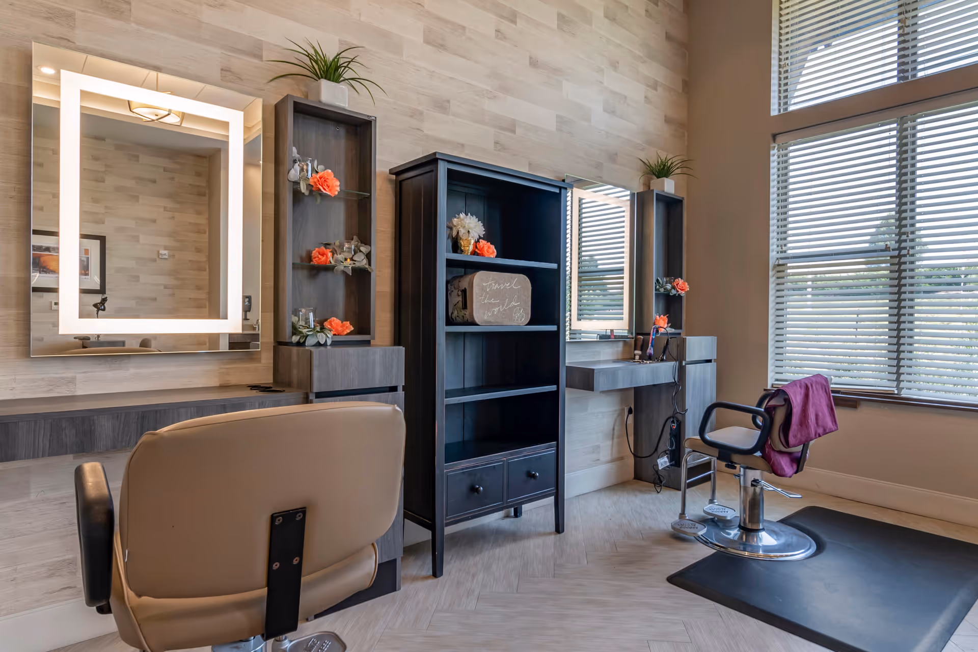 Salon-style interior with two styling chairs, illuminated mirrors, shelving with floral decor, and large windows with blinds.
