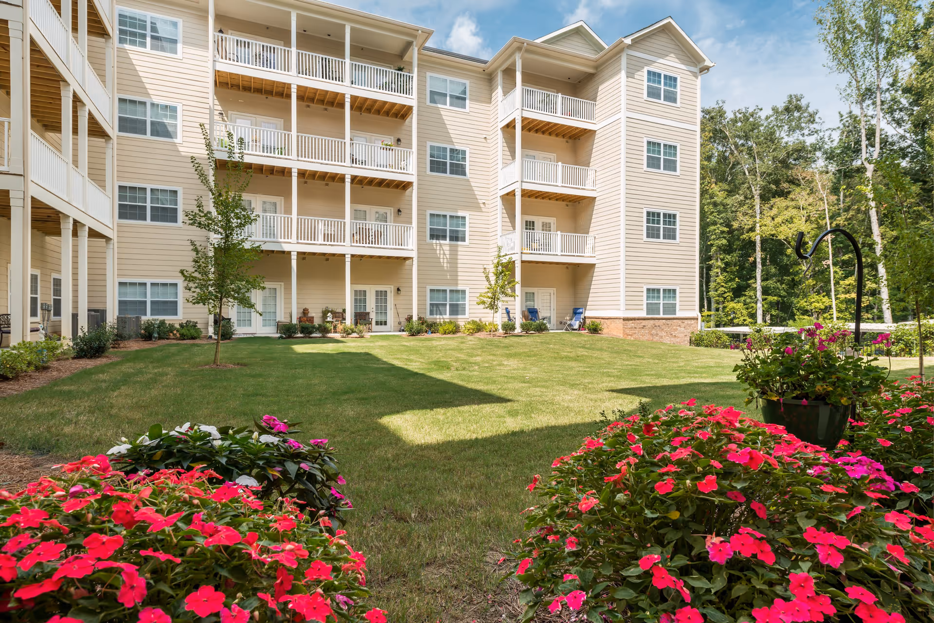 Exterior view of a beige multi-story senior living building with balconies overlooking a well-maintained grassy lawn and vibrant pink flowers in the foreground, surrounded by trees under a partly cloudy sky.