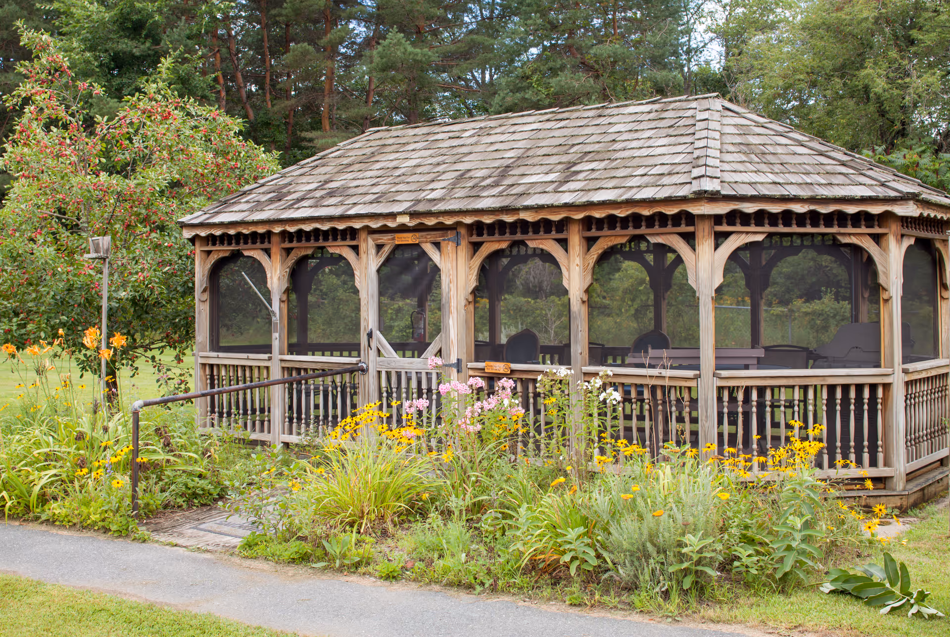A wooden gazebo with a shingled roof surrounded by colorful flowers and greenery in a garden setting. The gazebo has screened windows and contains several chairs inside. Trees and a grassy area are visible in the background.