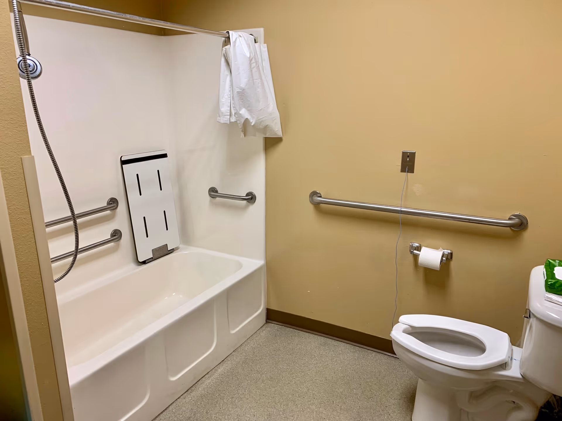 A bathroom with a white bathtub equipped with grab bars and a fold-down shower seat. There is a handheld showerhead attached to the wall. The toilet is next to the bathtub with a grab bar on the wall behind it and a toilet paper holder. The walls are painted beige and the floor has a light-colored speckled pattern.
