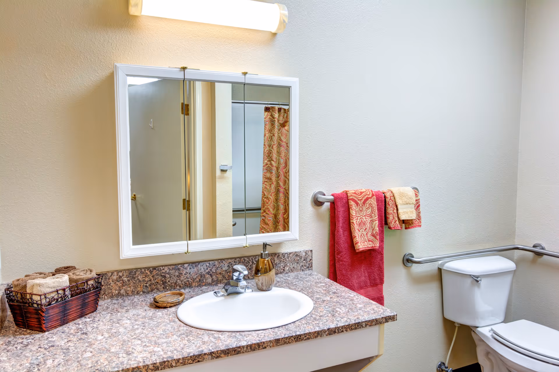 A clean bathroom featuring a white sink with a granite countertop, a wall-mounted mirror above the sink, a toilet with grab bars on both sides, and a towel rack holding red and patterned towels. A basket with rolled towels is placed on the countertop.