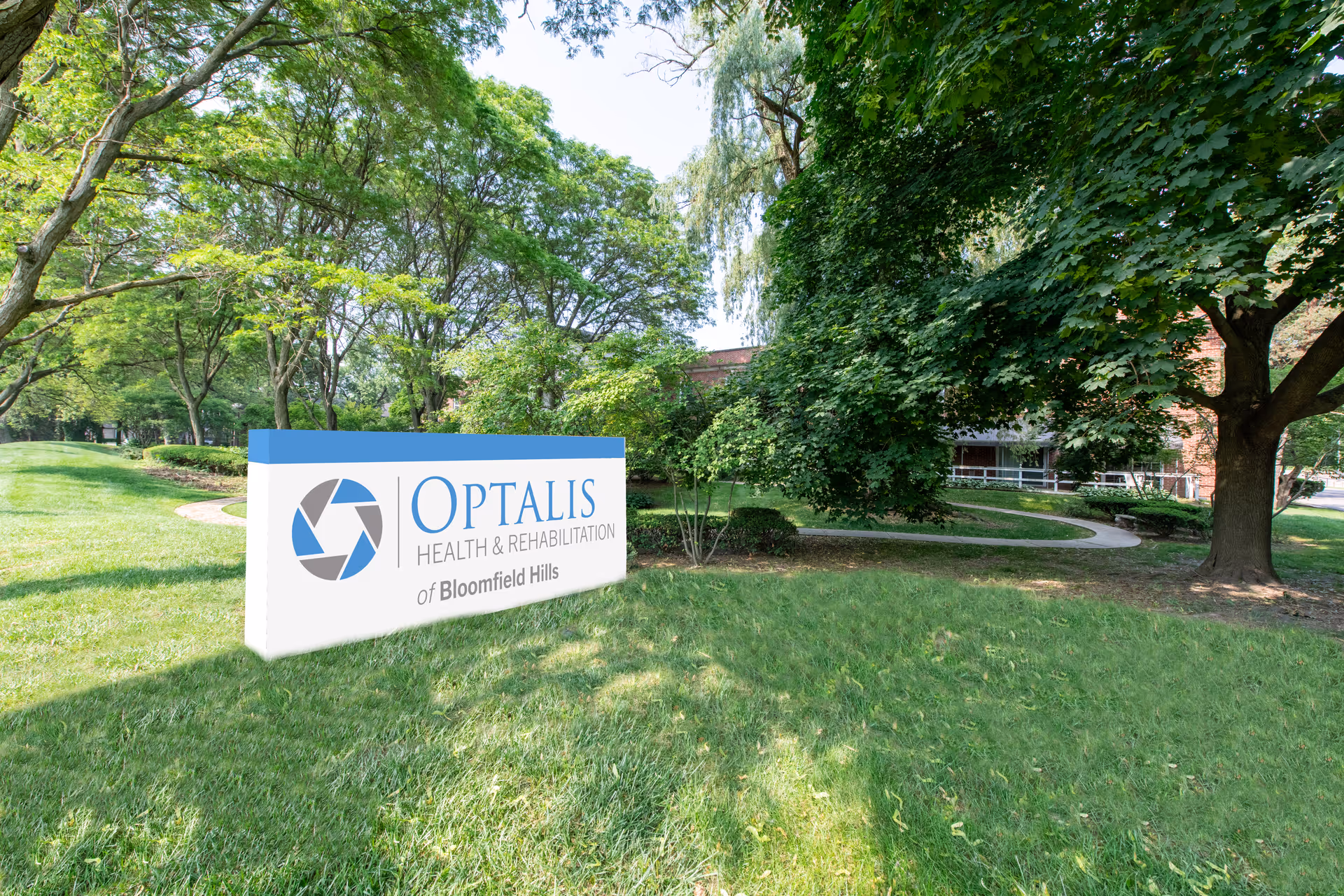 Sign for Optalis Health & Rehabilitation of Bloomfield Hills on a grassy lawn with trees and the facility building partially visible in the background.
