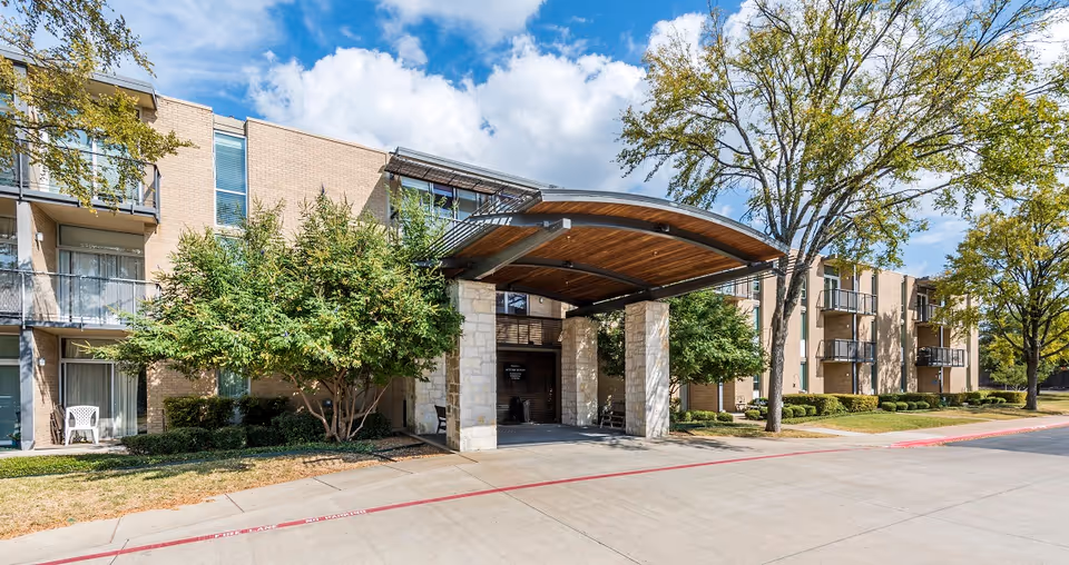Exterior view of a senior living facility named Autumn Leaves showing a three-story building with balconies, a covered entrance with stone pillars, trees, and a clear blue sky with some clouds.
