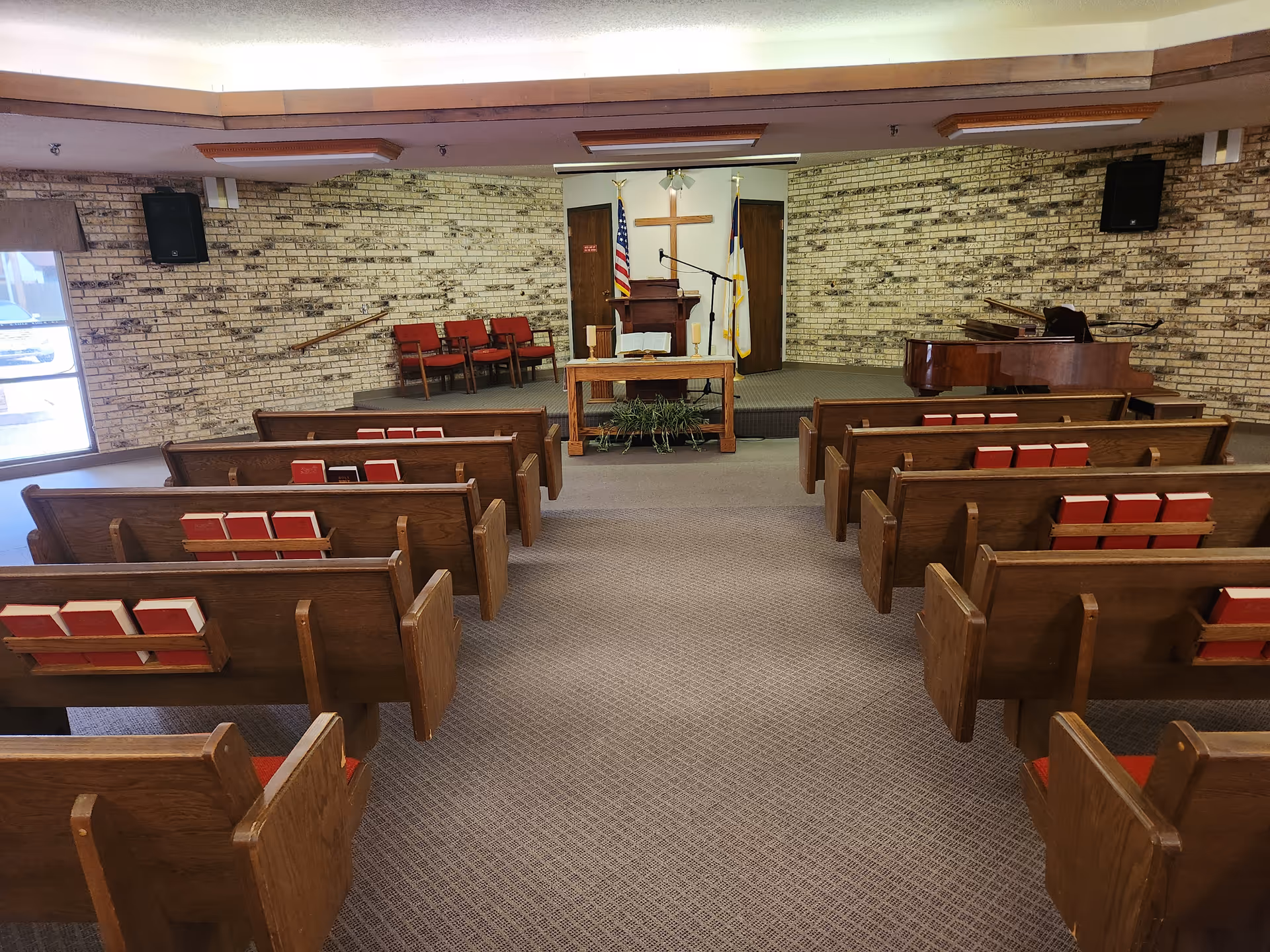 Interior view of a chapel or small church room with wooden pews arranged in rows facing a raised platform. On the platform, there is a wooden pulpit, a table with an open book, two candles, and a plant underneath. Behind the pulpit, there is a wooden cross mounted on a brick wall, flanked by an American flag and a Christian flag. To the right of the platform, there is a grand piano. The room has carpeted flooring and brick walls.