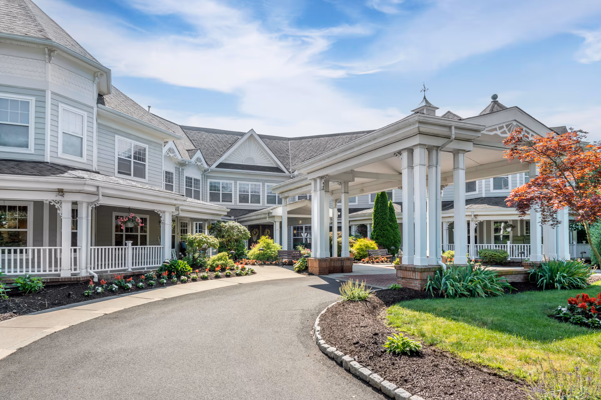 Exterior view of a senior living facility with a covered entrance, landscaped garden beds, and a paved driveway. The building has multiple windows, white railings, and a gray roof under a partly cloudy sky.