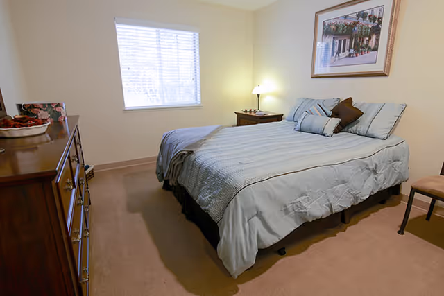 A neatly made bed with light blue bedding and several pillows in a bedroom. There is a wooden dresser on the left side with decorative items on top, a nightstand with a lamp on the right side of the bed, a window with blinds letting in natural light, and a framed picture hanging on the wall above the bed.
