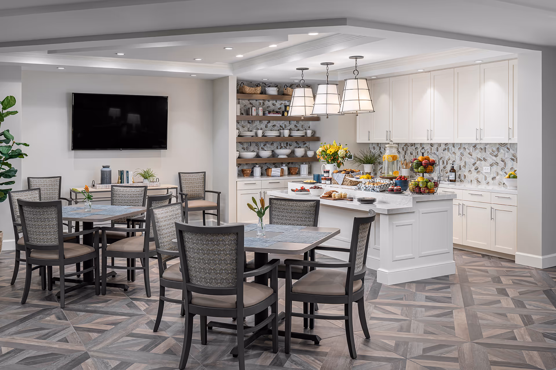 A modern dining area with several tables and chairs arranged on a patterned floor. The room features a kitchen area with white cabinets, open shelves with dishes, and a kitchen island topped with various foods and a large beverage dispenser. Three pendant lights hang above the island, and a flat-screen TV is mounted on the wall. There are small plants and flowers on the tables and counters, creating a welcoming atmosphere.