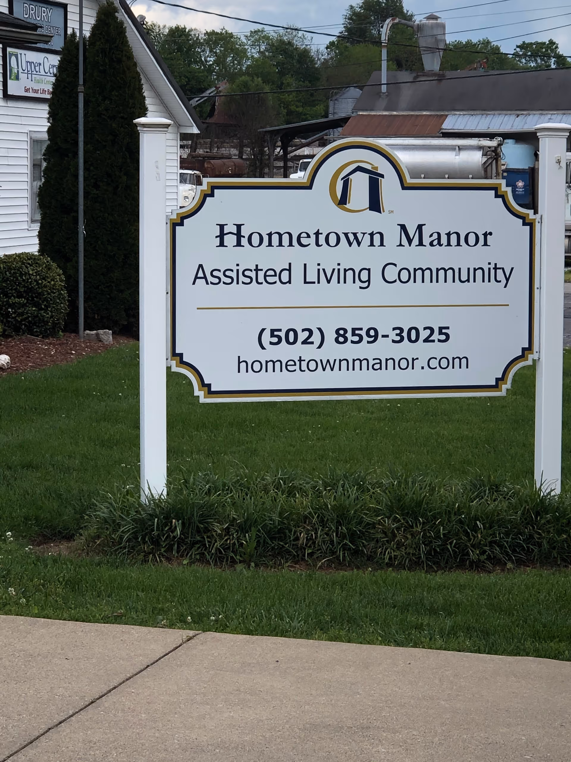 A white sign with decorative edges on a grassy lawn reads 'Hometown Manor Assisted Living Community' with a phone number and website below. The sign has a logo at the top featuring a stylized house. In the background, there is a white building, some bushes, and trees.