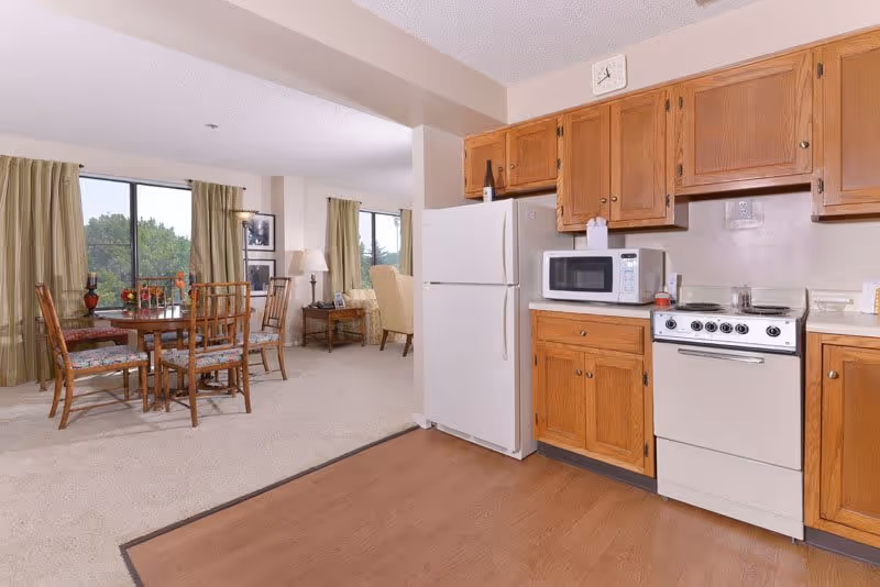 A bright kitchen area with wooden cabinets, a white refrigerator, microwave, and stove. Adjacent to the kitchen is a dining area with a round wooden table and four chairs with patterned cushions. Large windows with green curtains let in natural light, and a living area with armchairs and a side table is visible in the background.