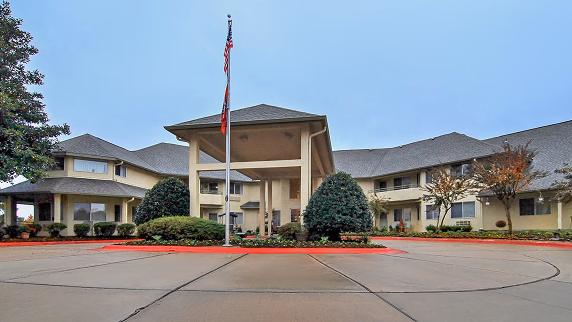 Front exterior view of Holiday Andover Place, a two-story senior living facility with a covered entrance, landscaped bushes, trees, and an American flag on a flagpole in front.