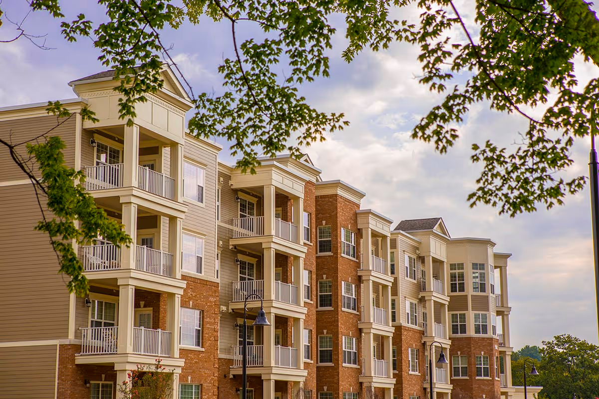 Exterior view of a multi-story residential building with balconies, beige and brick facade, and trees with green leaves in the foreground under a partly cloudy sky.