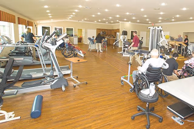 Spacious senior fitness room with treadmills, strength machines, and residents in wheelchairs using equipment under bright lighting.