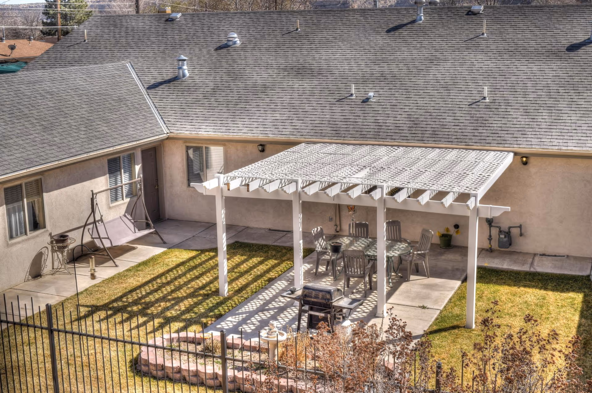 Outdoor courtyard area with a white pergola providing shade over a glass table and six chairs. There is a barbecue grill in front of the pergola, a swing bench near the building wall, and a small garden area with plants surrounded by a low brick border. The building has beige walls and a gray shingled roof.