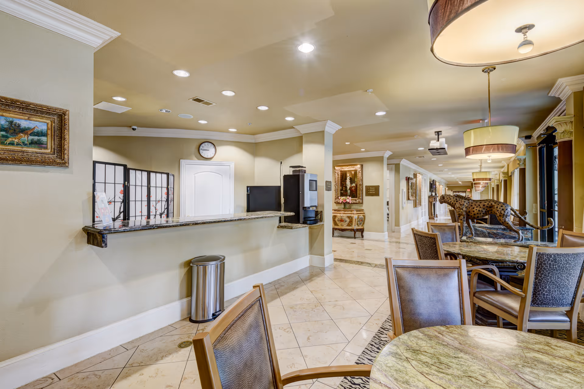 Interior view of a senior living facility featuring a seating area with round tables and chairs, a decorative statue of a cheetah on a table, pendant lights hanging from the ceiling, and a reception counter with a coffee machine and a clock on the wall behind it. The hallway extends into the background with framed artwork and decorative furniture along the walls.