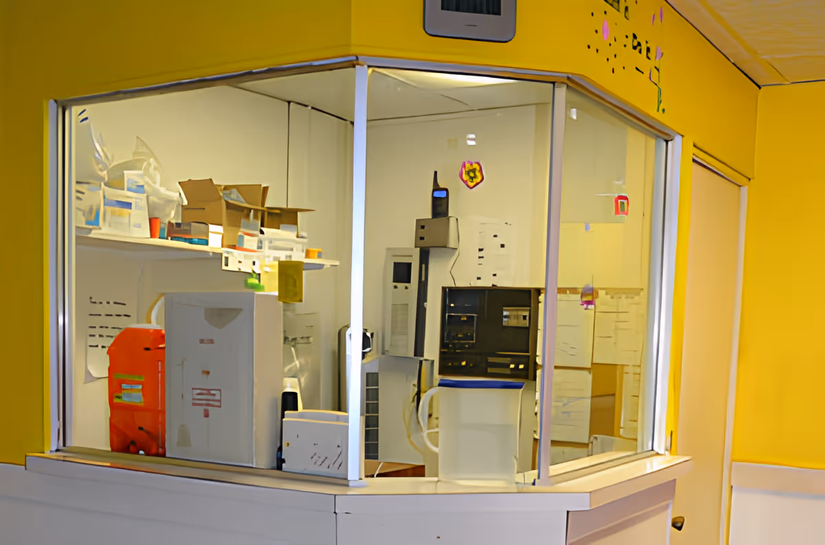 A small office or reception area with glass windows and a yellow wall. Inside, there are various boxes, papers, and office equipment including a computer and a walkie-talkie on top of a cabinet.