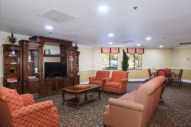 A cozy living room area with patterned carpet, a large wooden entertainment center with a TV, and several upholstered chairs and sofas in red and orange tones. There is a wooden coffee table in the center and a small dining table with chairs near the windows with red valances. Outside the windows, trees and greenery are visible.