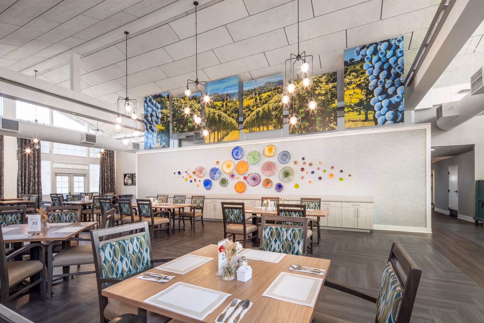 A bright and spacious dining room with multiple wooden tables and chairs featuring patterned upholstery. The room has large windows with curtains, modern hanging light fixtures, and colorful wall art including glass decorations and large photographic panels of vineyards and grapes.