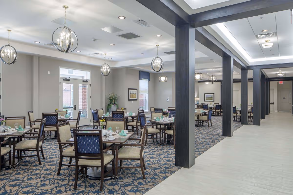 A spacious dining room in a senior living facility with multiple tables and chairs arranged neatly. The tables are set with plates, cups, and folded green napkins. The room features modern hanging light fixtures, patterned carpet, and large windows allowing natural light. Dark wooden beams separate the dining area from a light-colored tiled walkway.