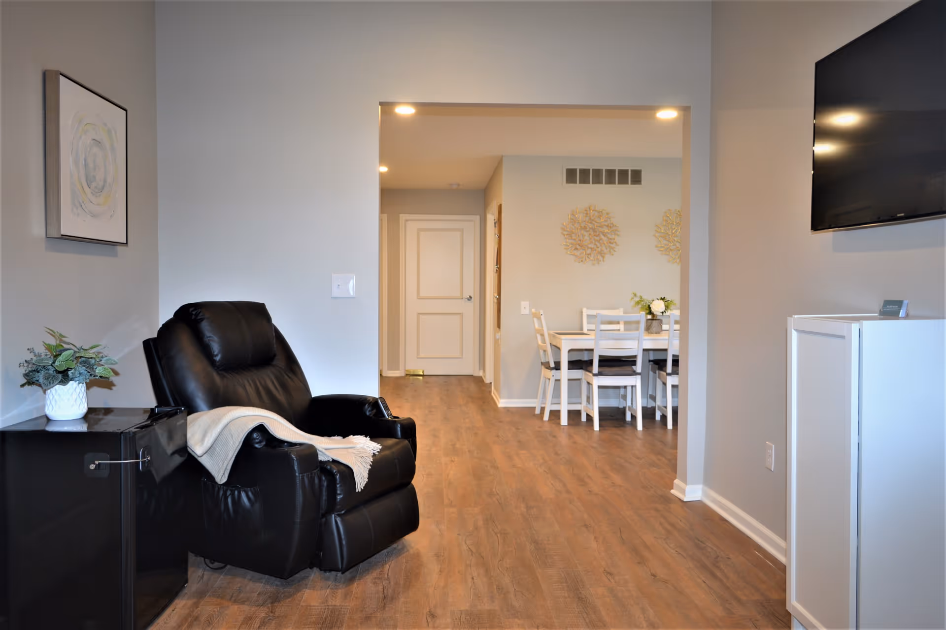 Interior view of a senior living facility showing a black leather recliner chair with a light-colored throw blanket draped over one arm, next to a small black side table with a potted plant. In the background, there is a dining area with a white dining table and chairs, decorated with a small flower arrangement and wall art. The floor is wooden, and the walls are painted light gray. A flat-screen TV is mounted on the wall to the right.