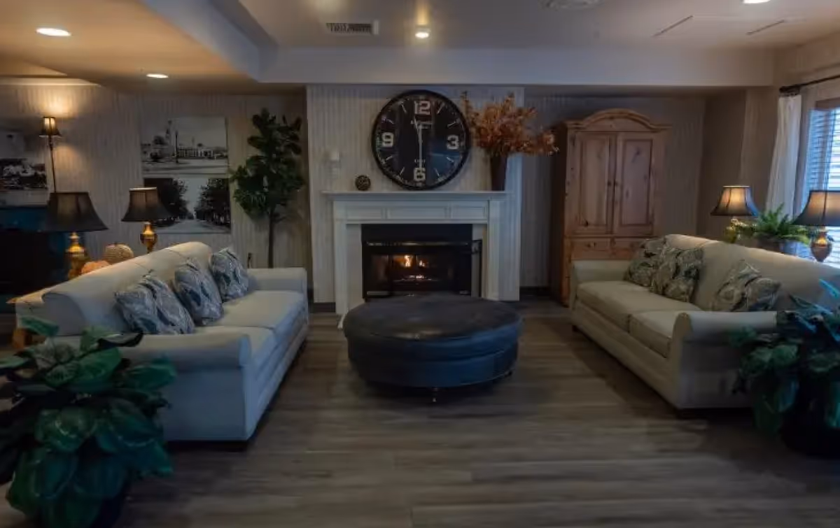 A cozy living-room style common area with two sofas facing a central ottoman and a fireplace topped by a large clock.