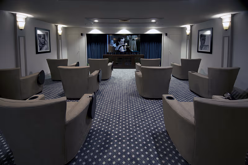 A small theater room with multiple gray upholstered armchairs arranged in rows facing a large flat-screen TV mounted on a wooden cabinet. The room has patterned carpet flooring, soft wall lighting, and framed black and white portraits on the walls.