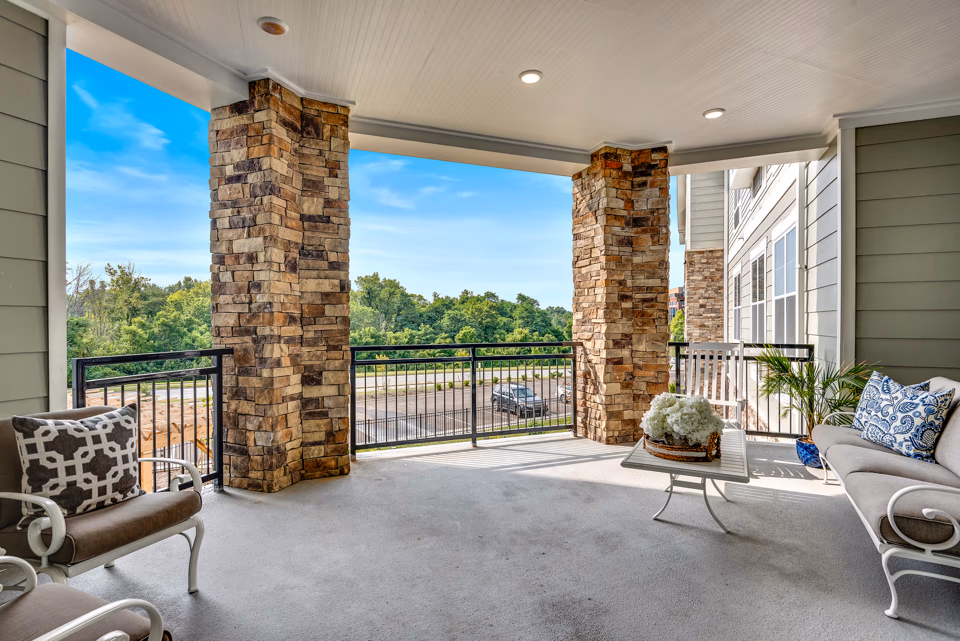 Covered outdoor patio area with stone pillars, metal railing, cushioned chairs, a small table with a flower arrangement, and a view of trees and a parking lot in the background under a blue sky.