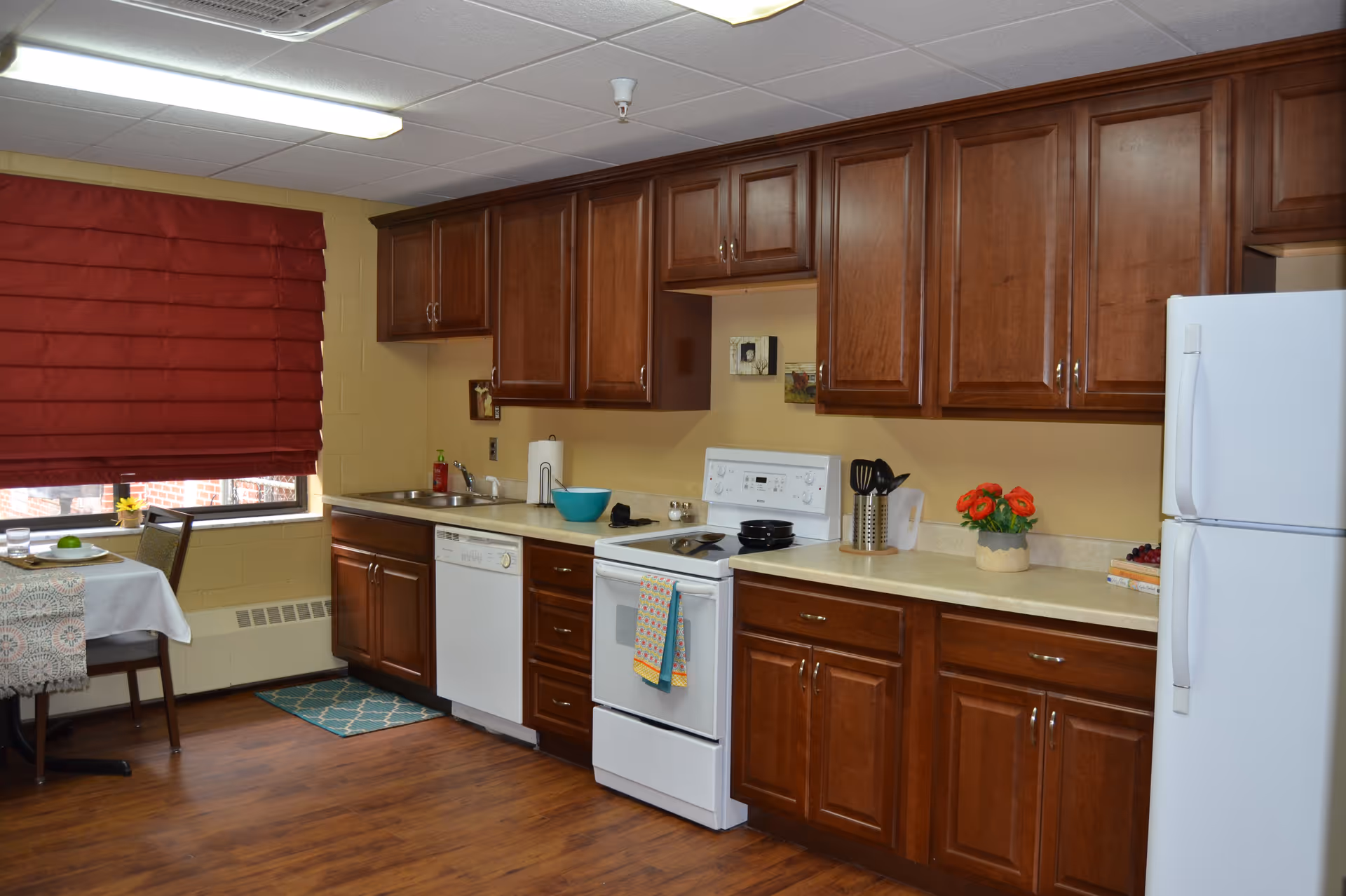 Interior kitchen with dark wood cabinets, white appliances (stove, refrigerator, dishwasher), a sink and countertop items, and a small table by a window with a red shade.