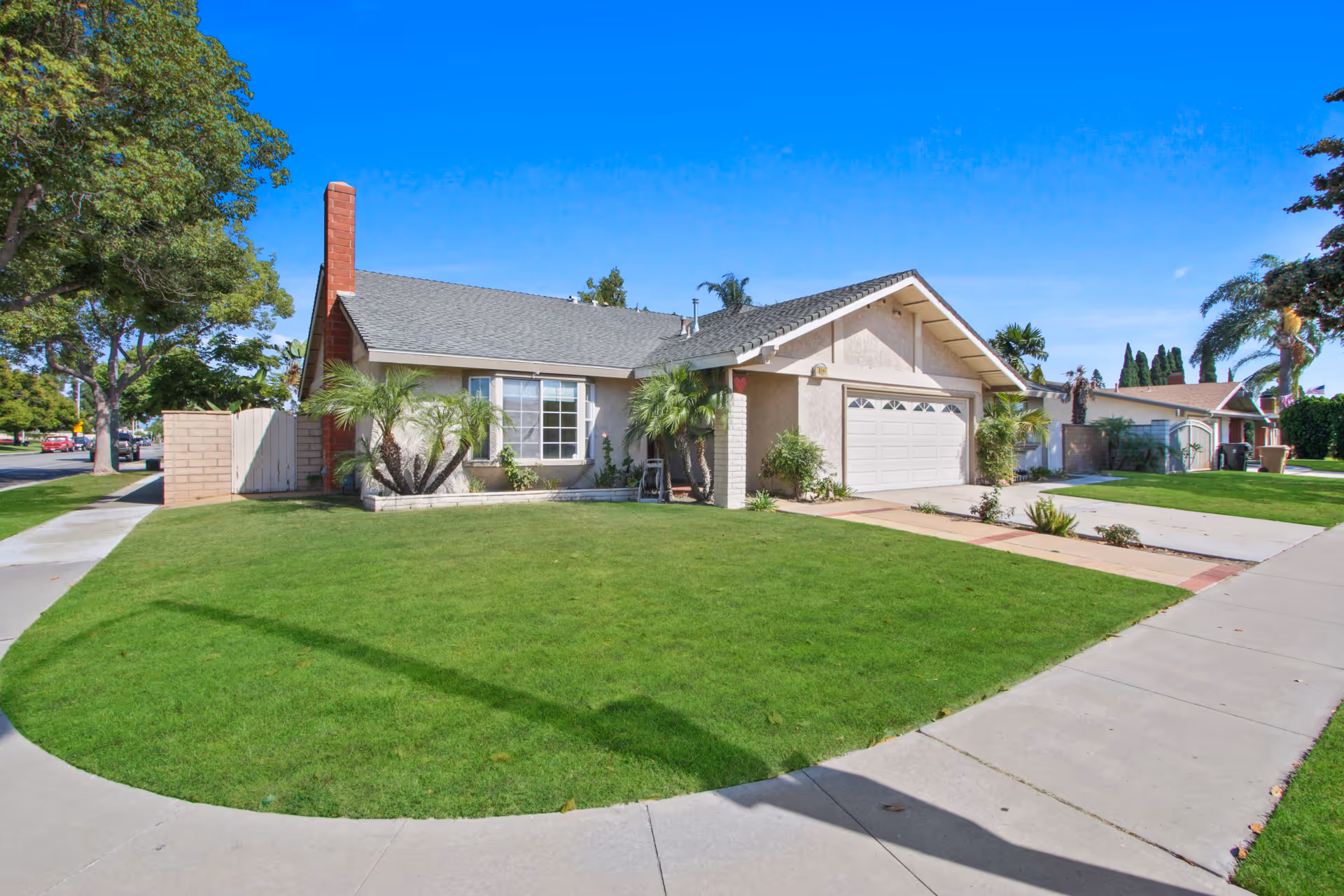 Single-story residential building with a gray shingled roof, beige exterior walls, and a two-car garage. The front yard has a well-maintained green lawn, several palm plants, and a concrete sidewalk curving around the property. Trees and neighboring houses are visible under a clear blue sky.