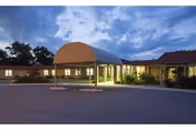 Exterior view of Legacy Heights Nursing Home at dusk, showing a covered entrance with a curved awning, well-lit building windows, and a paved driveway surrounded by landscaping and trees under a cloudy sky.