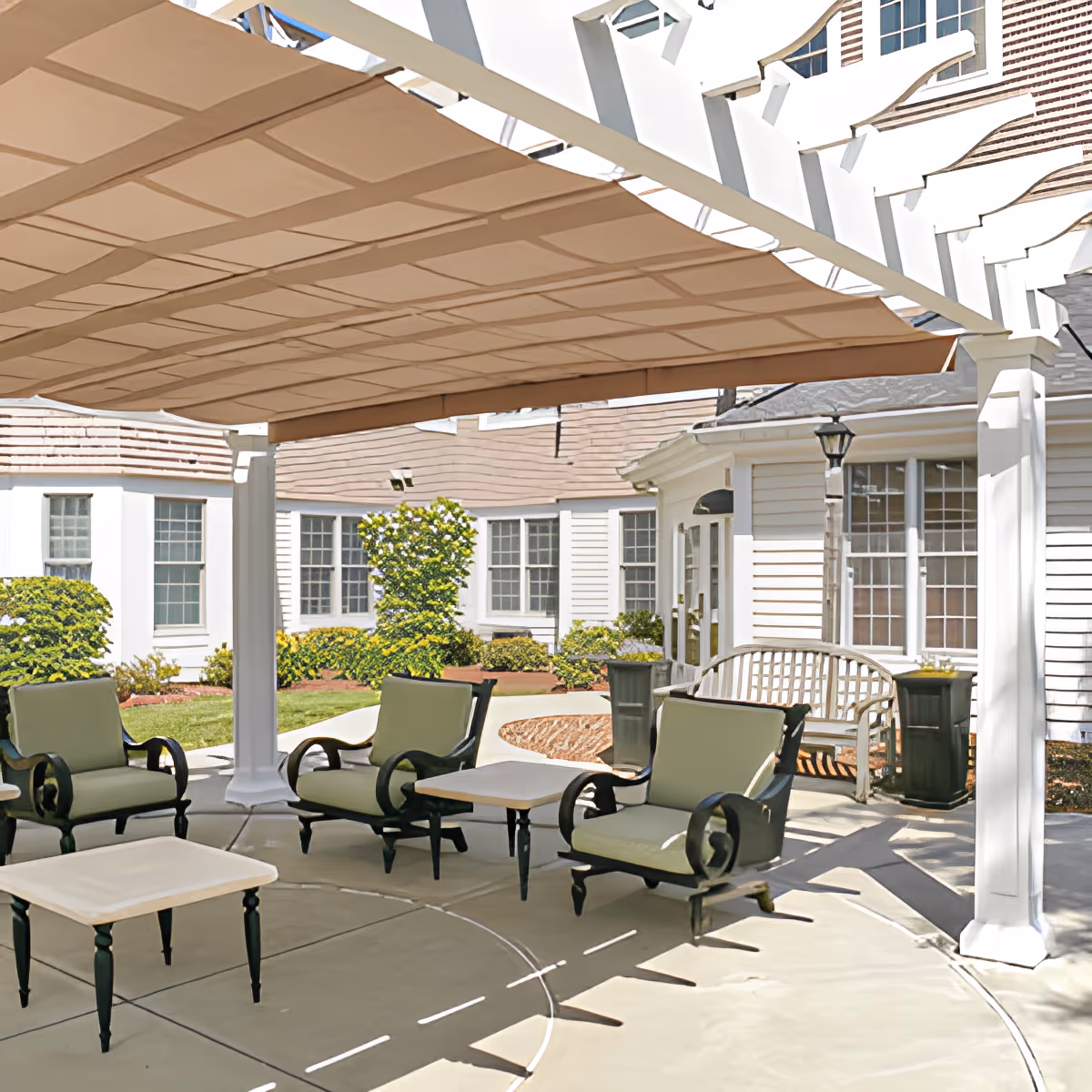 Outdoor patio area with cushioned chairs and tables under a pergola with a fabric canopy, surrounded by white buildings with multiple windows and some greenery.