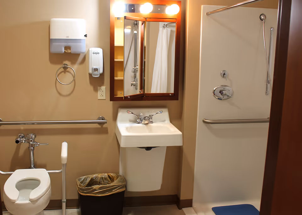 A bathroom in Good Samaritan Home featuring a toilet with support bars, a sink with a mirror cabinet above it, a shower area with a grab bar and a shower curtain, a paper towel dispenser, a soap dispenser, and a trash bin.