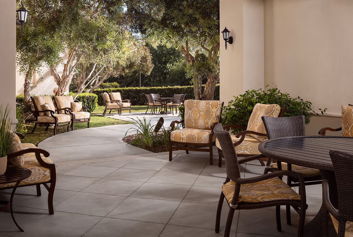 Outdoor patio area with cushioned chairs and tables surrounded by greenery and trees, featuring a tiled floor and wall-mounted lanterns.