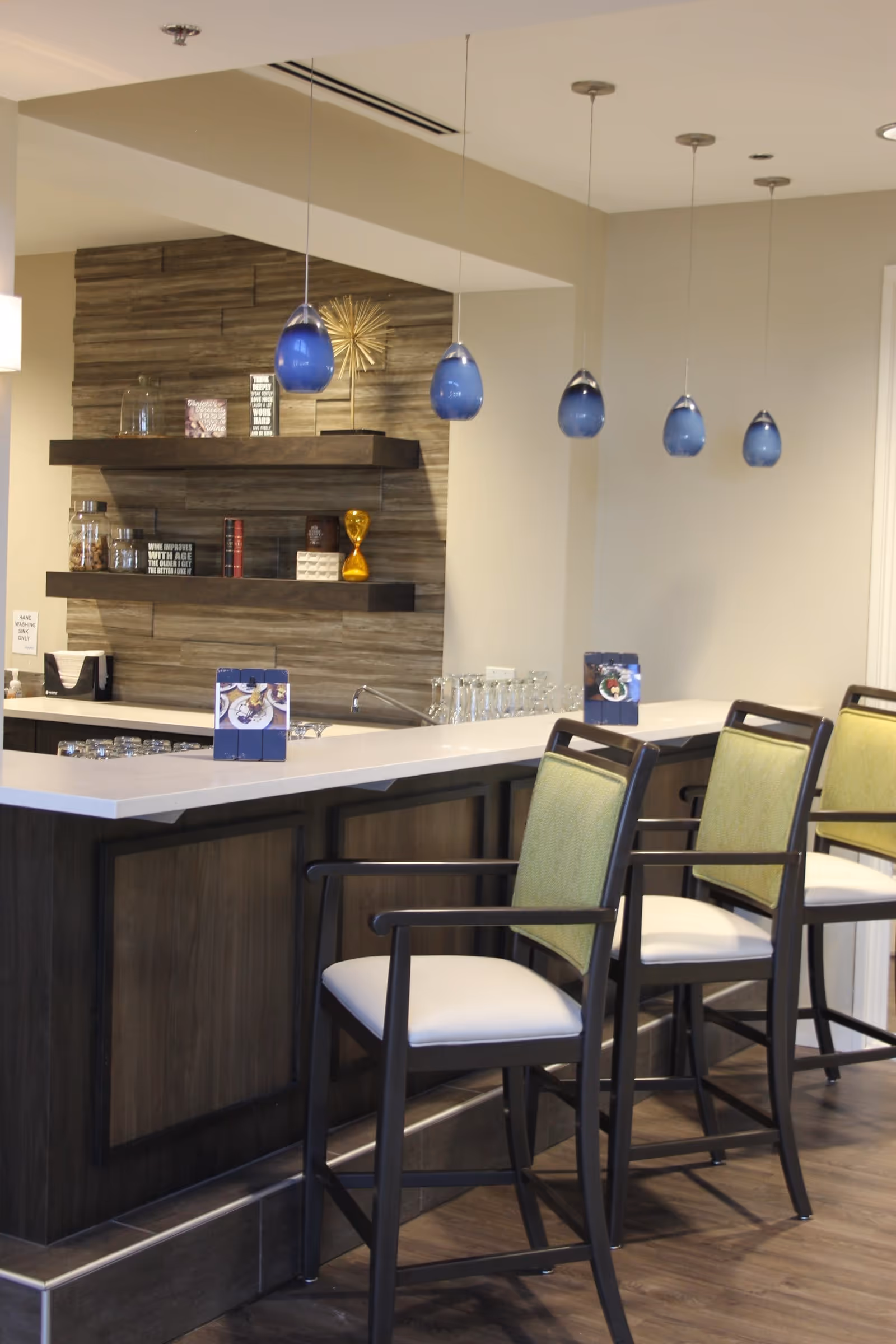 Interior view of a modern bar area with a white countertop and dark wood paneling. Four high chairs with green backs and white seats are lined up along the bar. Above the bar hang five blue pendant lights. Behind the bar, there are two floating shelves with decorative items including jars, books, and a golden hourglass. The floor is wooden, and the walls are painted light beige.