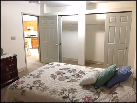 A bedroom with a bed covered in a floral quilt and several pillows. There is a wooden dresser on the left side and two open closets with double doors on the far wall. A doorway leads to a kitchen area with wooden cabinets and appliances visible.