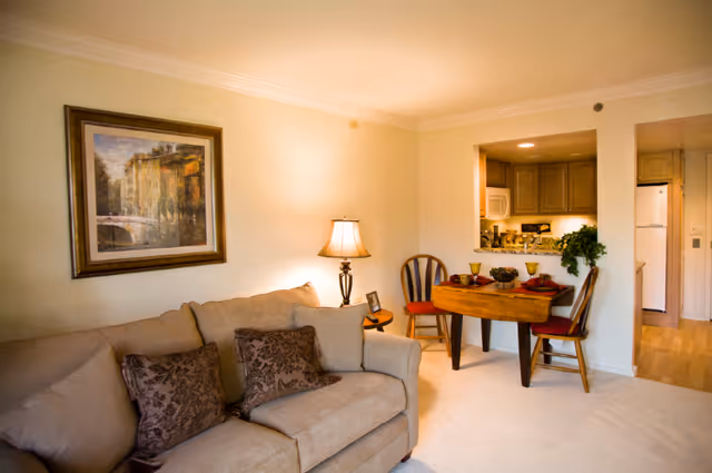 A cozy living room area with a beige couch adorned with patterned pillows, a side table with a lamp, and a framed painting on the wall. Adjacent to the living room is a small dining area with a wooden table set for two with red placemats and chairs. The kitchen is visible through a pass-through window, featuring cabinets, a microwave, and a refrigerator.