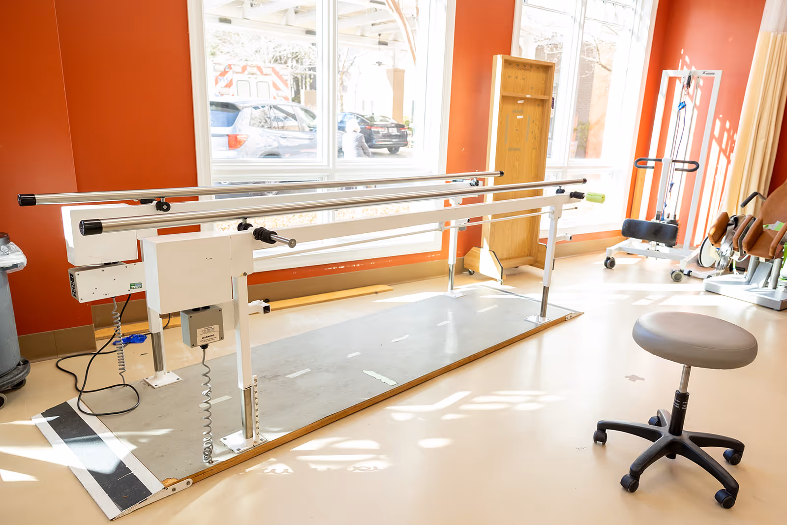 A bright room with large windows showing parallel bars used for physical therapy or rehabilitation exercises. There is a gray rolling stool in the foreground and various exercise equipment in the background. The walls are painted orange and the floor is light-colored.