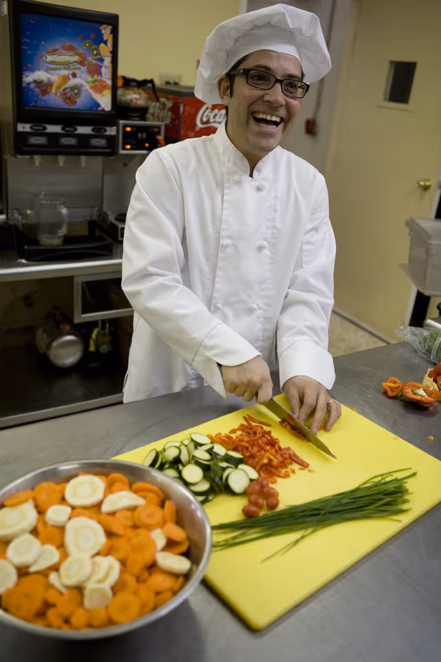 A smiling chef wearing a white chef's coat and hat is chopping red bell peppers on a yellow cutting board in a kitchen. There are sliced cucumbers, cherry tomatoes, and chives also on the cutting board, with a bowl of sliced carrots and parsnips nearby. Kitchen equipment and a soda machine are visible in the background.