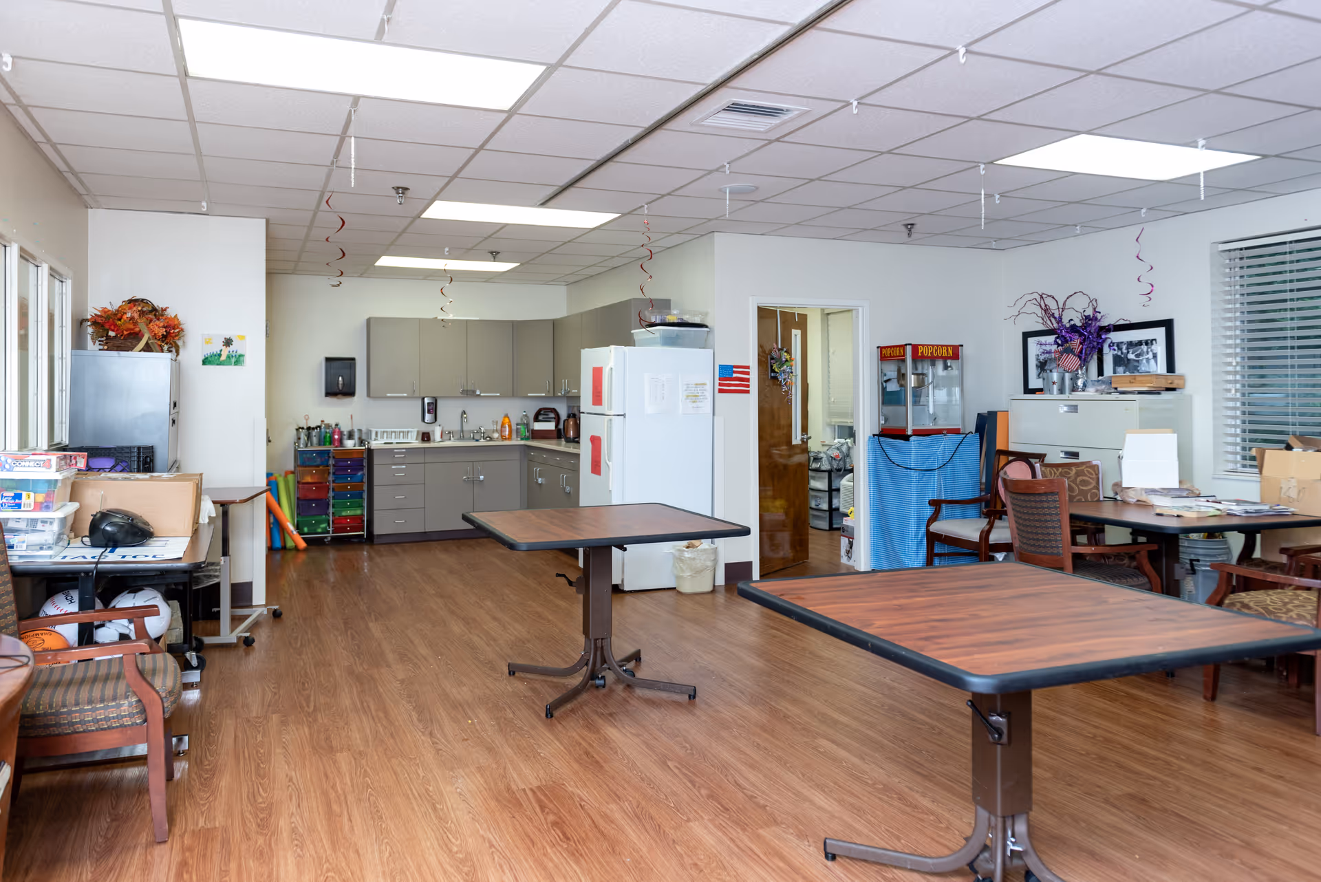 Interior view of a multipurpose room with a kitchenette in the background, featuring gray cabinets, a white refrigerator, and a sink. The room has wood flooring, two square tables with dark edges, chairs, a popcorn machine, and various office supplies and decorations including flowers and wall art.