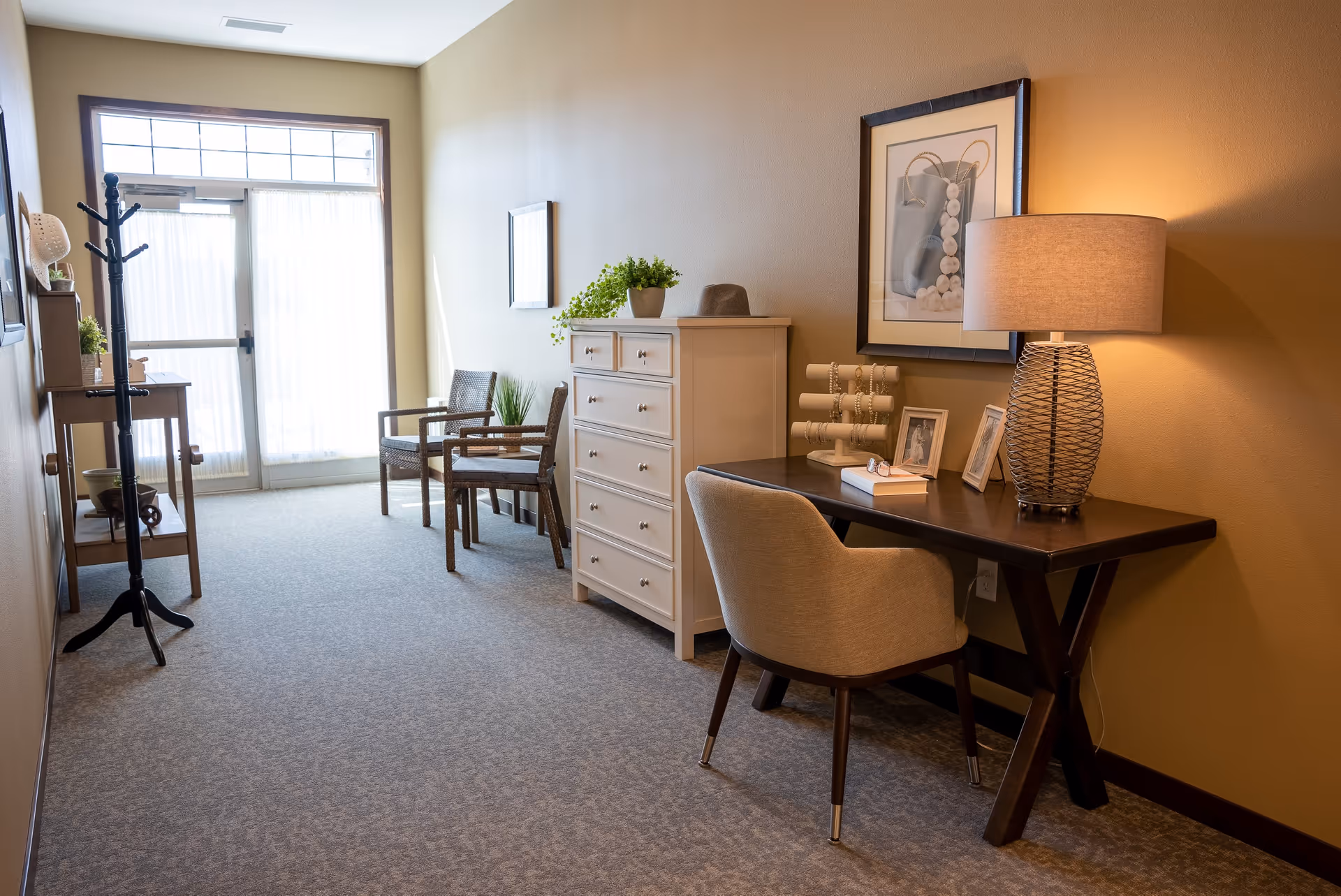 A well-lit interior hallway in a senior living facility with a desk and chair on the right side, a lamp, framed pictures, and a jewelry stand on the desk. There is a white chest of drawers next to the desk, two wicker chairs with a small table between them near a large window and glass door letting in natural light. A coat rack and a small table with decorative items are on the left side of the hallway.