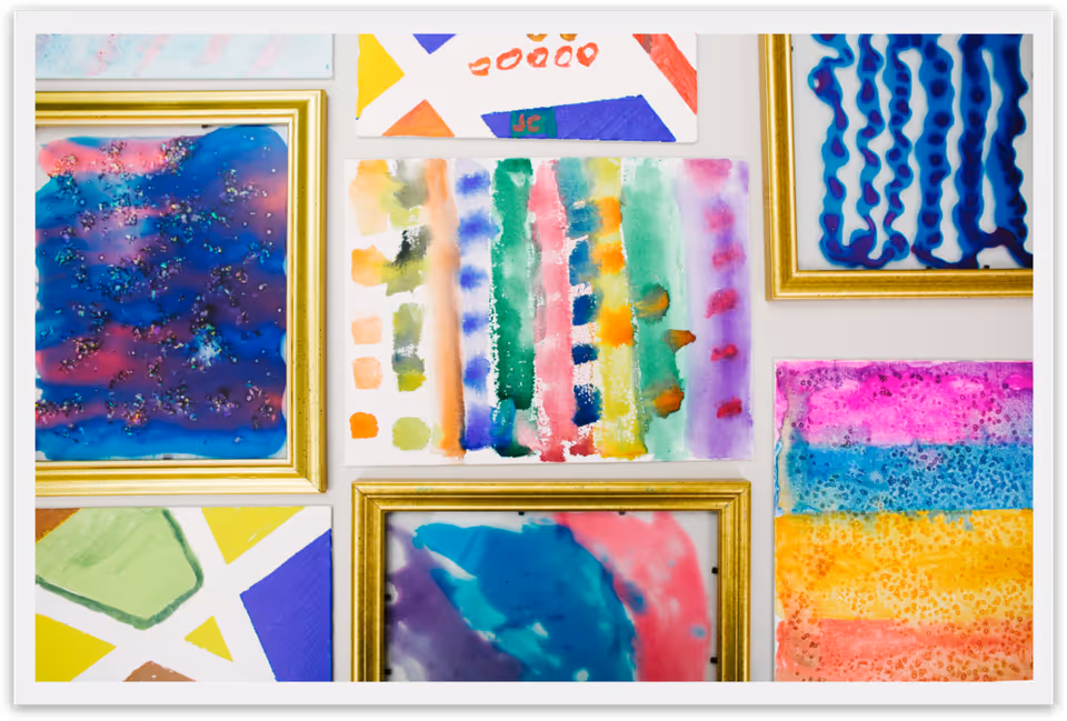 Colorful abstract watercolor paintings and prints in gold frames arranged on a white wall.