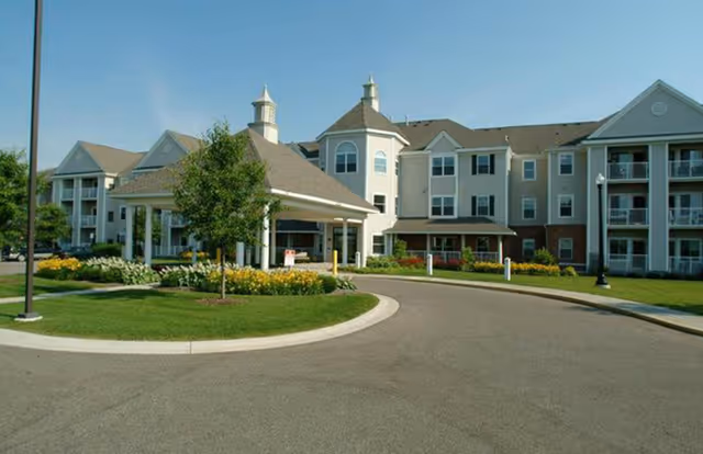 Front exterior of a multi-story senior living building with a covered porte-cochère, circular driveway, and landscaped grounds.