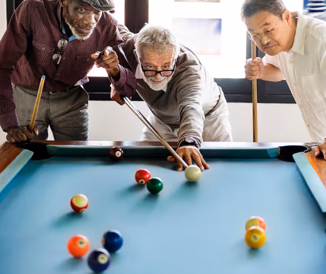 Three elderly men playing pool together indoors, one man is aiming to hit the cue ball with a pool cue while the other two watch closely.
