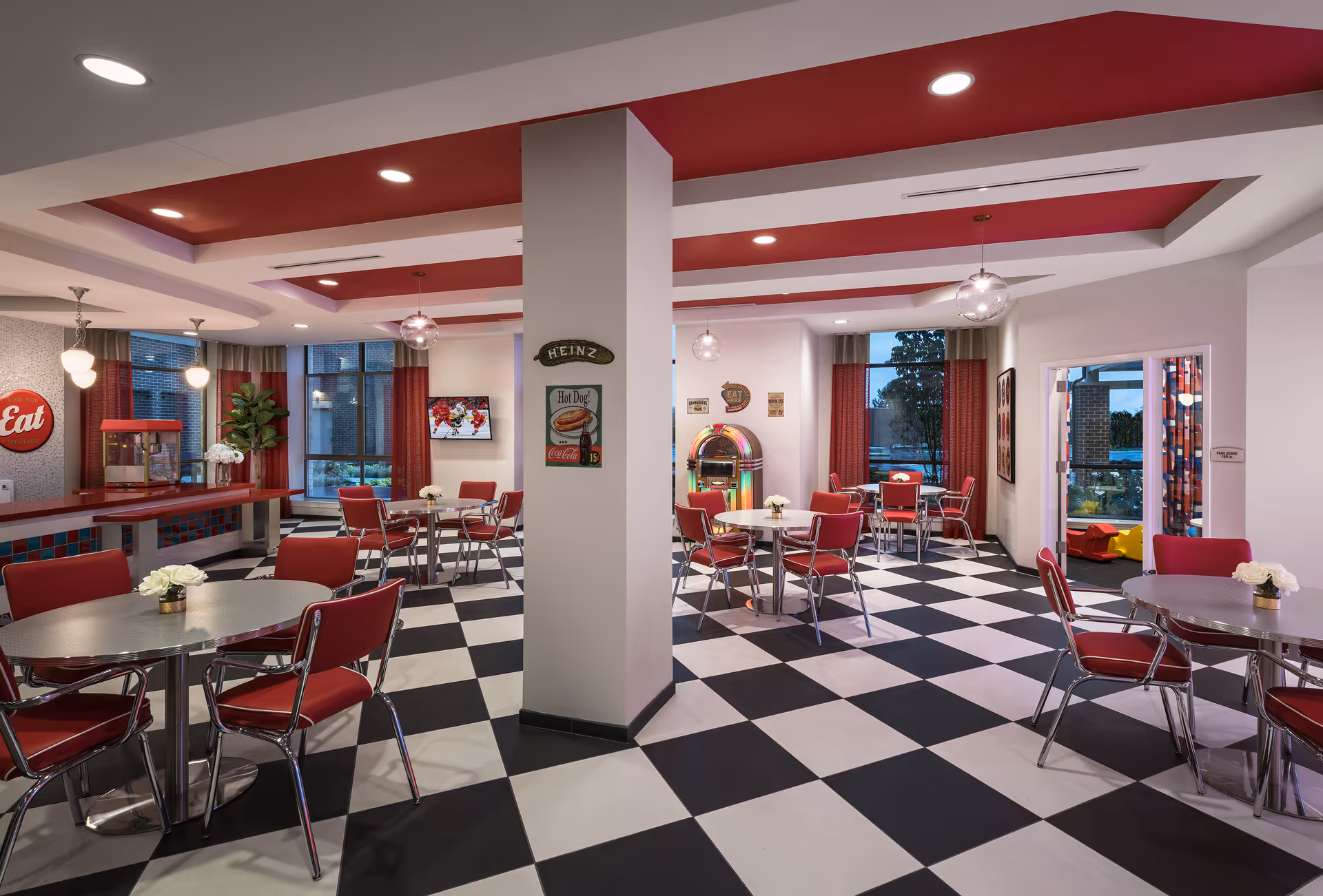 A retro-style dining room with black and white checkered flooring, red and chrome chairs around round tables, red ceiling accents, and pendant lights. The room features a jukebox, a popcorn machine, and vintage-style wall decorations including a Heinz hot dog sign and an 'Eat' sign. Large windows with red curtains allow natural light into the space.