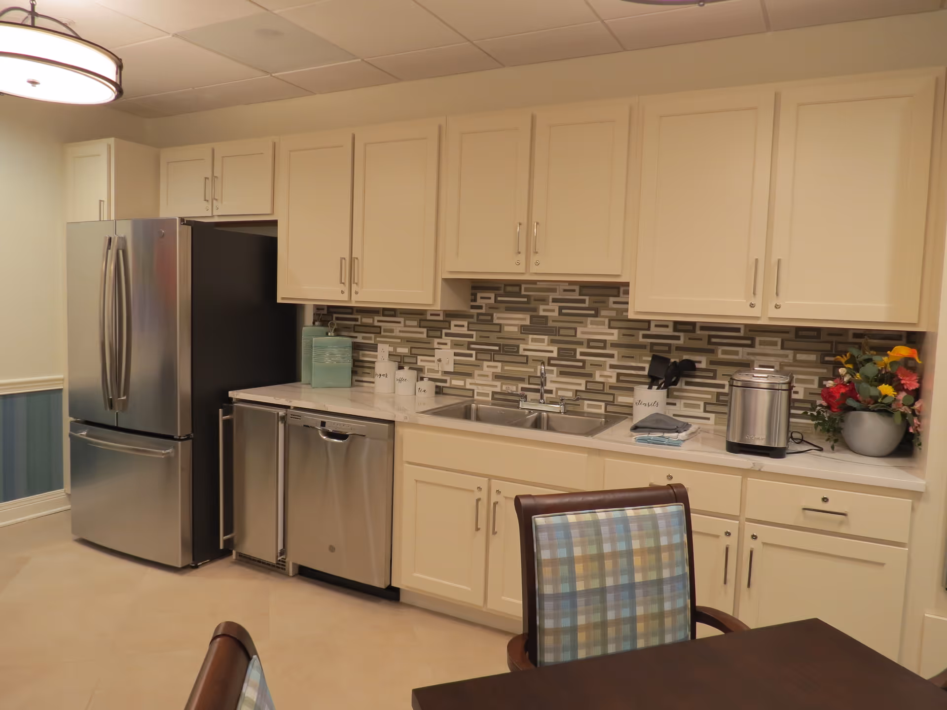 A modern kitchen area with cream-colored cabinets, a stainless steel refrigerator, dishwasher, and a double sink. The backsplash features a mosaic tile design in shades of gray and beige. On the countertop, there are decorative containers, a utensil holder, a small appliance, and a flower arrangement. In the foreground, part of a dining table and a chair with a plaid upholstered back are visible.
