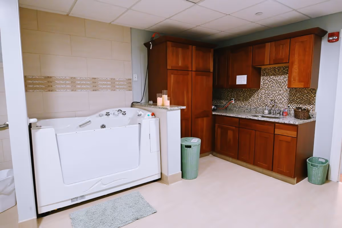 A bathroom area featuring a white walk-in bathtub with control knobs and a safety handle, adjacent to a wooden cabinet and countertop with a sink. The walls have beige tiles with a decorative horizontal strip, and the floor is light-colored. There are two green trash bins and some candles on the countertop.