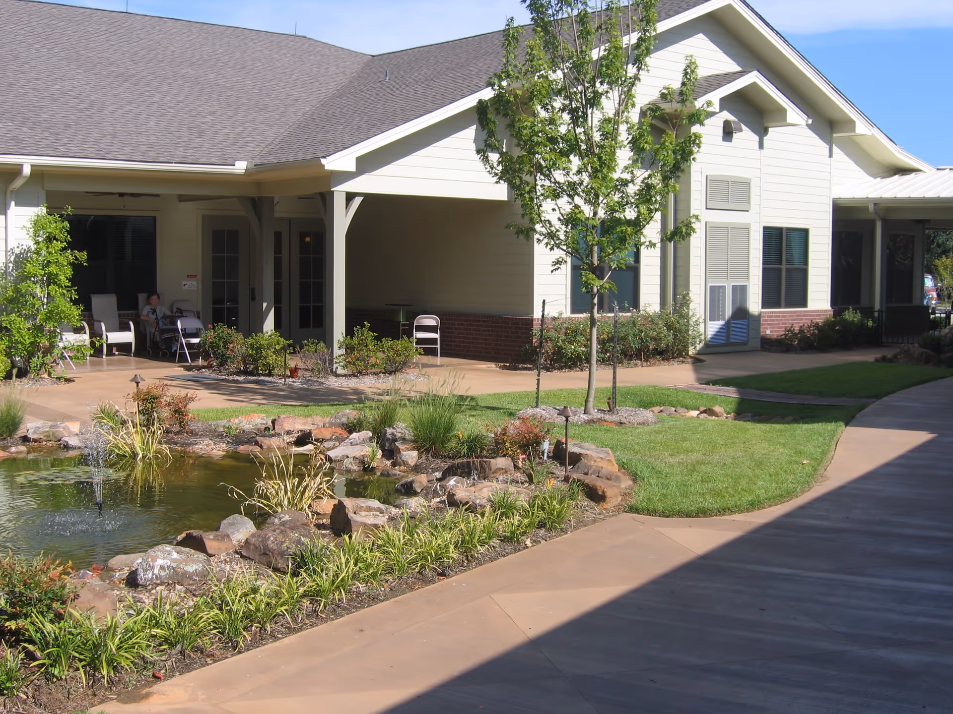 Outdoor view of a senior living facility with a small landscaped pond surrounded by rocks and plants, a paved walkway, green grass, and a building with a covered porch area featuring chairs and a person sitting.