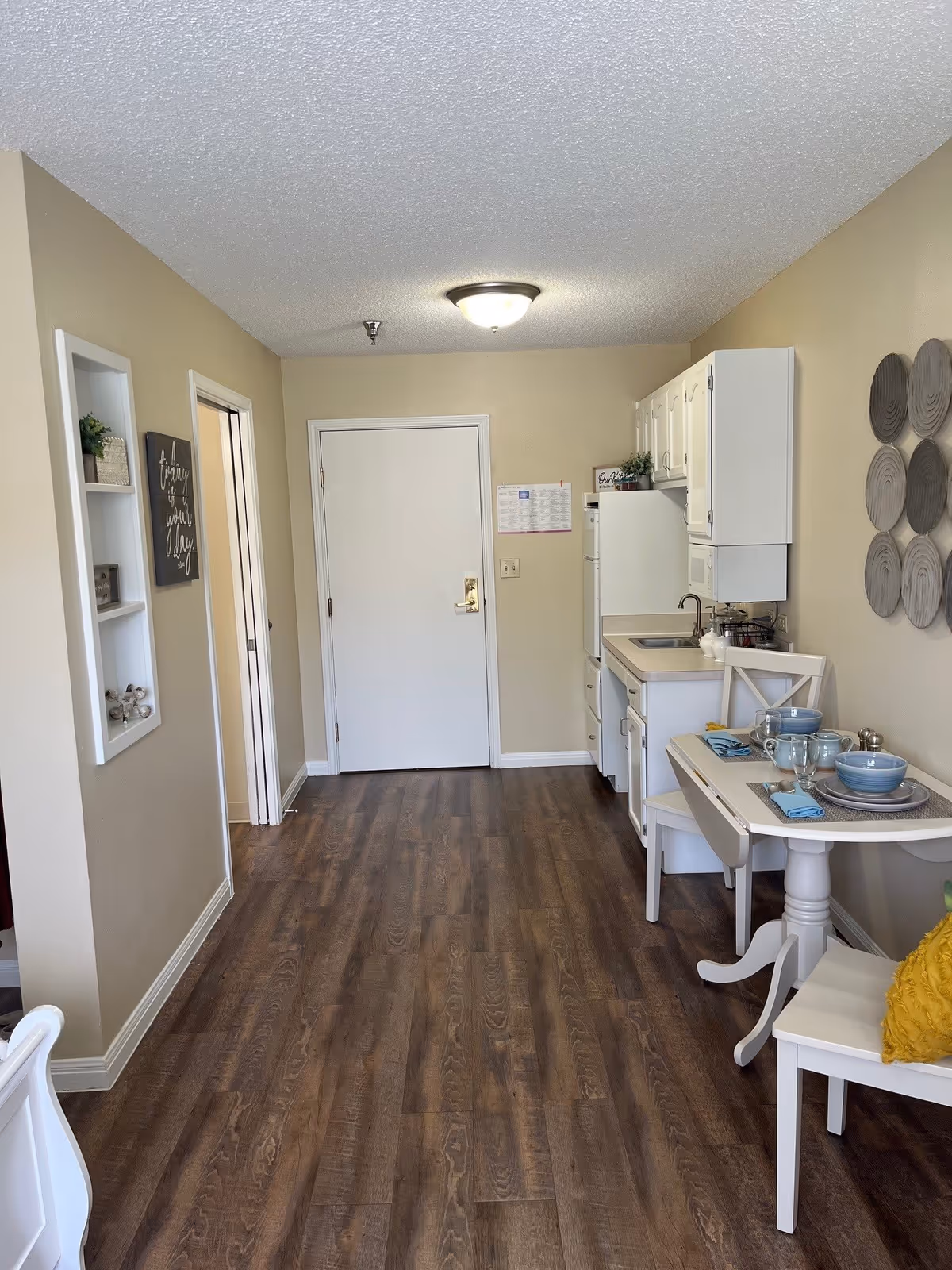 Interior view of a small kitchen and dining area in a senior living facility. The room has wood flooring, beige walls, and a white ceiling with a ceiling light. On the right side, there is a compact kitchen with white cabinets, a sink, a refrigerator, and a small dining table set for two with blue dishes and glasses. Decorative wall art is visible above the dining table. On the left wall, there is a small recessed shelf with decorative items and a framed sign. A closed white door is at the end of the room.